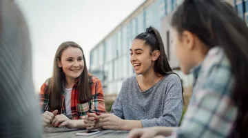 Three girls sit at a table outside of school talking and smiling